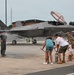 A military pilot in a flight suit is being sprayed with water by family members on an airfield tarmac in front of an F-35 Lightning II fighter jet bearing tail number 5816. This appears to be a traditional 'fini flight' or final flight celebration, a military aviation tradition where a pilot completing their last flight is doused with water. A small group of family members, including children, and military personnel in the background are participating in the ceremony.