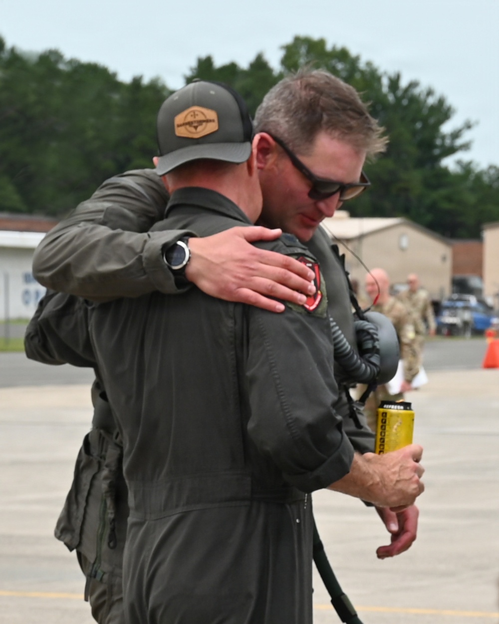 Two individuals in olive/sage green flight suits are embracing on what appears to be a military airfield tarmac. One person is wearing a gray baseball cap with a leather patch logo, while the other is wearing sunglasses and holding what appears to be a yellow can. Military personnel in camouflage uniforms are visible in the background.