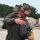 104th Fighter Wing Celebrates Flight Surgeon's Fifteen Years of Service with Fini Flight Two individuals in olive/sage green flight suits are embracing on what appears to be a military airfield tarmac. One person is wearing a gray baseball cap with a leather patch logo, while the other is wearing sunglasses and holding what appears to be a yellow can. Military personnel in camouflage uniforms are visible in the background.
