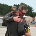 Two individuals in olive/sage green flight suits are embracing on what appears to be a military airfield tarmac. One person is wearing a gray baseball cap with a leather patch logo, while the other is wearing sunglasses and holding what appears to be a yellow can. Military personnel in camouflage uniforms are visible in the background.
