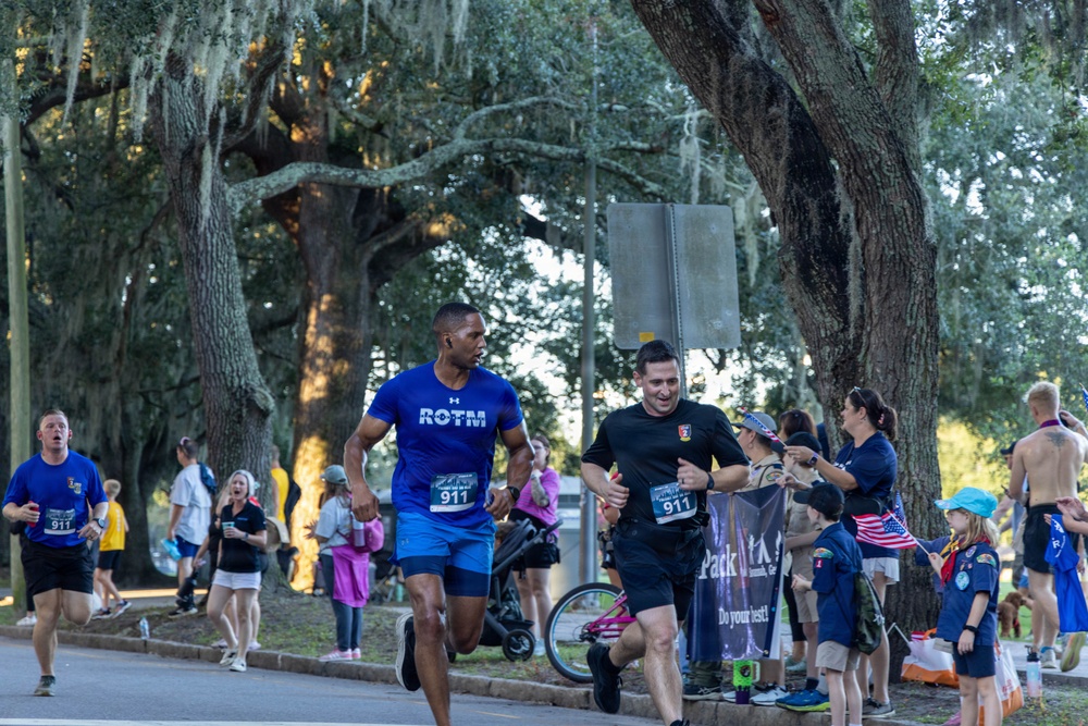 A road race event is underway, with multiple runners wearing bib number 911 passing through a cheering crowd. The scene appears to take place along a tree-lined street with Spanish moss-draped oak trees, suggesting a Southern U.S. location. Spectators including Boy Scouts and families line the course, holding signs and cheering on the participants.