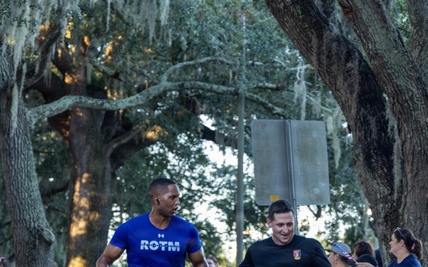 A road race event is underway, with multiple runners wearing bib number 911 passing through a cheering crowd. The scene appears to take place along a tree-lined street with Spanish moss-draped oak trees, suggesting a Southern U.S. location. Spectators including Boy Scouts and families line the course, holding signs and cheering on the participants.