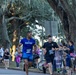 A road race event is underway, with multiple runners wearing bib number 911 passing through a cheering crowd. The scene appears to take place along a tree-lined street with Spanish moss-draped oak trees, suggesting a Southern U.S. location. Spectators including Boy Scouts and families line the course, holding signs and cheering on the participants.