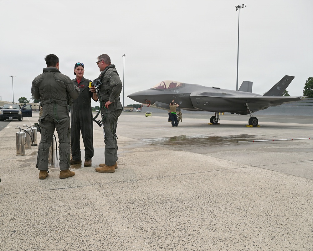 Three individuals in military flight suits stand on a wet aircraft ramp, engaged in conversation. In the background, an F-35 Lightning II stealth fighter jet is parked on the tarmac with ground crew personnel nearby. The setting appears to be a military air base under overcast conditions.