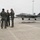 104th Fighter Wing Celebrates Flight Surgeon's Fifteen Years of Service with Fini Flight Three individuals in military flight suits stand on a wet aircraft ramp, engaged in conversation. In the background, an F-35 Lightning II stealth fighter jet is parked on the tarmac with ground crew personnel nearby. The setting appears to be a military air base under overcast conditions.