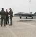 Three individuals in military flight suits stand on a wet aircraft ramp, engaged in conversation. In the background, an F-35 Lightning II stealth fighter jet is parked on the tarmac with ground crew personnel nearby. The setting appears to be a military air base under overcast conditions.