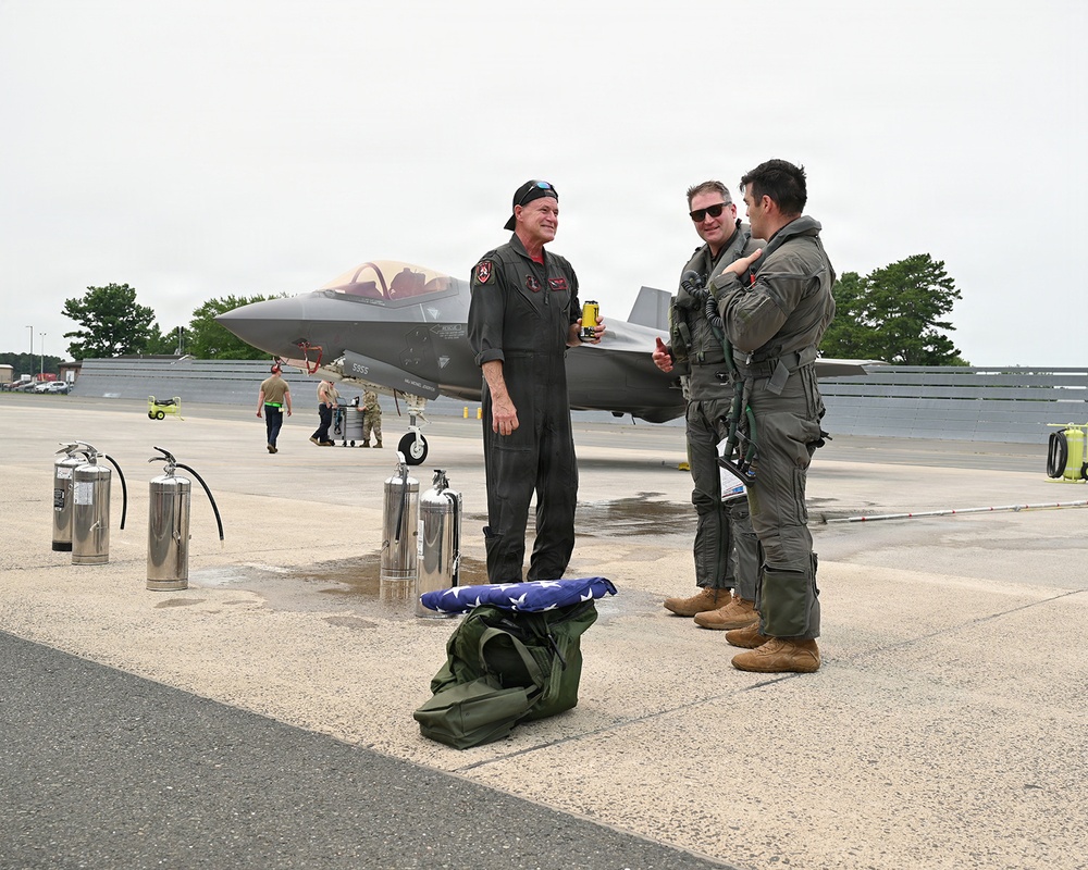 Three military aviators in flight suits are conversing on an airfield tarmac in front of what appears to be an F-35 Lightning II stealth fighter jet. A folded American flag rests on a green duffel bag in the foreground, suggesting a ceremonial occasion such as a final flight or retirement. Additional personnel and ground equipment are visible in the background.