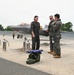 Three military aviators in flight suits are conversing on an airfield tarmac in front of what appears to be an F-35 Lightning II stealth fighter jet. A folded American flag rests on a green duffel bag in the foreground, suggesting a ceremonial occasion such as a final flight or retirement. Additional personnel and ground equipment are visible in the background.