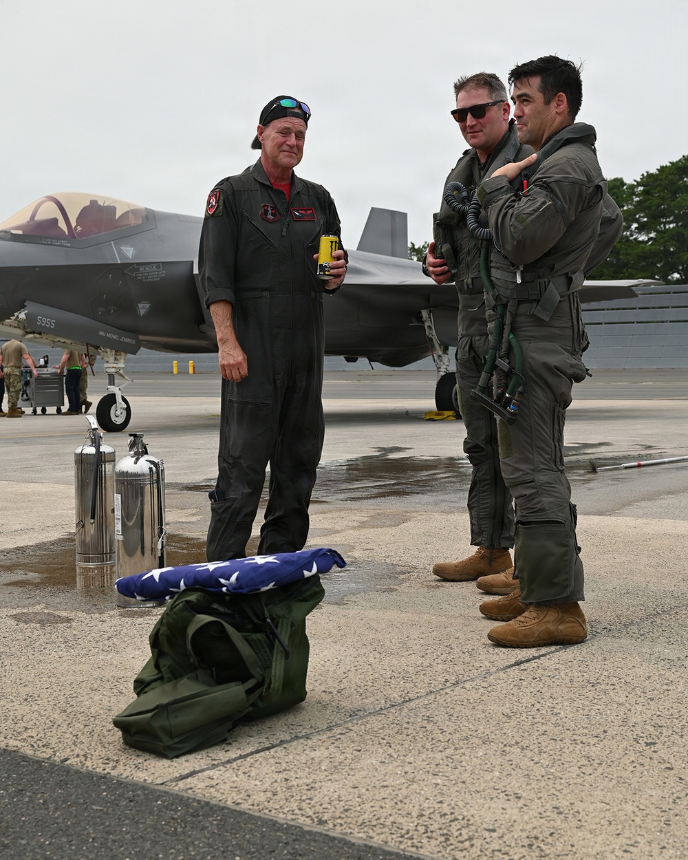 Three military personnel in flight suits stand on a wet aircraft tarmac near an F-35 fighter jet. A folded American flag rests on top of a green duffel bag in the foreground, suggesting a ceremonial occasion such as a retirement flight or memorial event. The scene appears to be at a military air base with ground support equipment visible in the background.