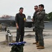 Three military personnel in flight suits stand on a wet aircraft tarmac near an F-35 fighter jet. A folded American flag rests on top of a green duffel bag in the foreground, suggesting a ceremonial occasion such as a retirement flight or memorial event. The scene appears to be at a military air base with ground support equipment visible in the background.
