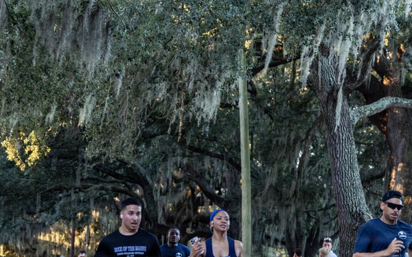 A road race event is taking place on a tree-lined street canopied with large live oak trees draped in Spanish moss, suggesting a Southern U.S. location such as Georgia or Florida. Multiple runners wearing bib number 911 are prominently visible in the foreground, with spectators lining the sidewalk. The lighting suggests early morning or late afternoon conditions.