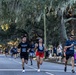 A road race event is taking place on a tree-lined street canopied with large live oak trees draped in Spanish moss, suggesting a Southern U.S. location such as Georgia or Florida. Multiple runners wearing bib number 911 are prominently visible in the foreground, with spectators lining the sidewalk. The lighting suggests early morning or late afternoon conditions.