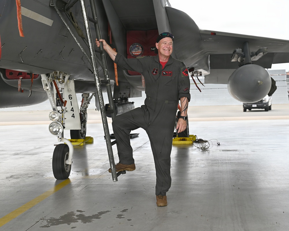 A smiling male aviator in an olive/gray flight suit stands beside a military jet aircraft on a flight line or tarmac. He is posing with one foot on the boarding ladder and one hand gripping it, appearing relaxed and confident. The aircraft appears to be an F-15 based on its general shape and markings.