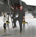 A smiling male aviator in an olive/gray flight suit stands beside a military jet aircraft on a flight line or tarmac. He is posing with one foot on the boarding ladder and one hand gripping it, appearing relaxed and confident. The aircraft appears to be an F-15 based on its general shape and markings.