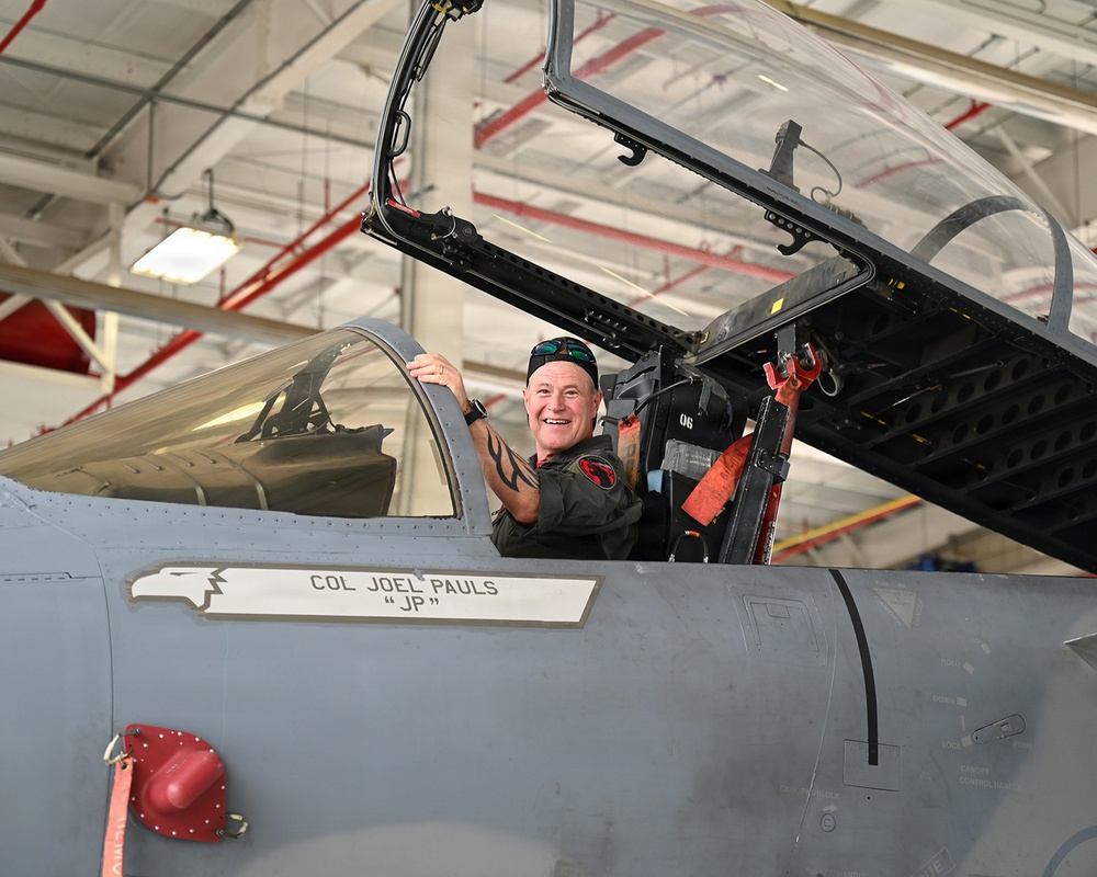 A smiling military pilot, identified as Col. Joel Pauls with the callsign 'JP', is seated in the cockpit of a gray military jet fighter inside a hangar. The aircraft's canopy is open and the pilot is wearing a dark flight suit with a squadron patch. The name placard on the fuselage clearly identifies the assigned pilot.