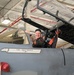 A smiling military pilot, identified as Col. Joel Pauls with the callsign 'JP', is seated in the cockpit of a gray military jet fighter inside a hangar. The aircraft's canopy is open and the pilot is wearing a dark flight suit with a squadron patch. The name placard on the fuselage clearly identifies the assigned pilot.
