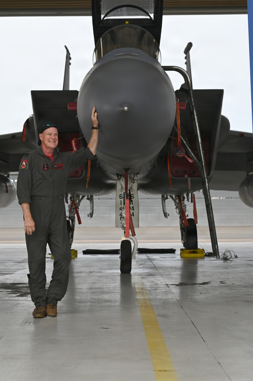 A smiling male aviator in an olive/gray flight suit stands at the nose of a military jet aircraft inside a hangar. He is posing with one hand resting on the large radome (nose cone) of what appears to be an F-15 Eagle. The aircraft is on chocks with safety streamers and ground equipment visible.