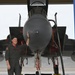 A smiling male aviator in an olive/gray flight suit stands at the nose of a military jet aircraft inside a hangar. He is posing with one hand resting on the large radome (nose cone) of what appears to be an F-15 Eagle. The aircraft is on chocks with safety streamers and ground equipment visible.