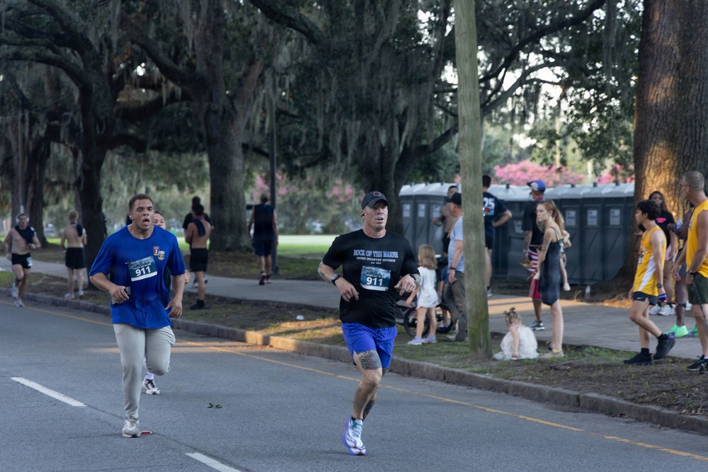 Two runners wearing matching bib number 911 are competing in an outdoor road race along a tree-lined street. The setting appears to be a organized running event, likely a 5K or similar race, with spectators and portable toilets visible in the background. The race takes place beneath large oak trees draped with Spanish moss, suggesting a Southern U.S. location such as Savannah, Georgia.