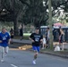 Two runners wearing matching bib number 911 are competing in an outdoor road race along a tree-lined street. The setting appears to be a organized running event, likely a 5K or similar race, with spectators and portable toilets visible in the background. The race takes place beneath large oak trees draped with Spanish moss, suggesting a Southern U.S. location such as Savannah, Georgia.