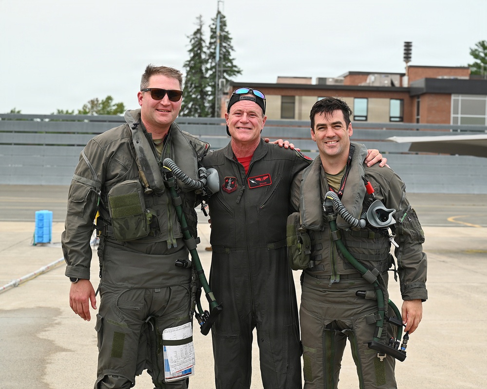 Three men pose together on what appears to be a military airfield tarmac. Two of the men are wearing full flight suits with survival gear, oxygen masks, and harnesses, while the center individual wears a black flight suit with a red shirt and unit patches. All three are smiling, suggesting a celebratory or commemorative occasion.