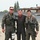 104th Fighter Wing Celebrates Flight Surgeon's Fifteen Years of Service with Fini Flight Three men pose together on what appears to be a military airfield tarmac. Two of the men are wearing full flight suits with survival gear, oxygen masks, and harnesses, while the center individual wears a black flight suit with a red shirt and unit patches. All three are smiling, suggesting a celebratory or commemorative occasion.