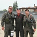 Three men pose together on what appears to be a military airfield tarmac. Two of the men are wearing full flight suits with survival gear, oxygen masks, and harnesses, while the center individual wears a black flight suit with a red shirt and unit patches. All three are smiling, suggesting a celebratory or commemorative occasion.