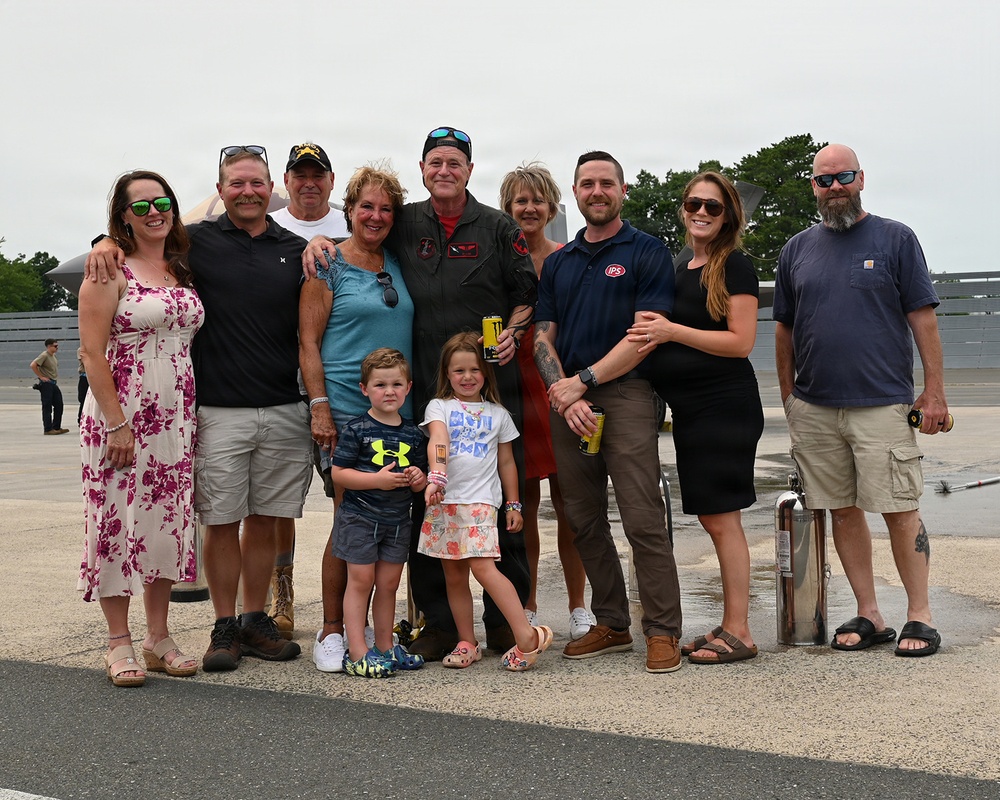 A group of approximately 12 people, including two young children, pose together on a military airfield tarmac. A man in a military flight suit stands at the center of the group, suggesting this may be a retirement ceremony, final flight, or homecoming event. A military aircraft is partially visible in the background.
