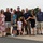 104th Fighter Wing Celebrates Flight Surgeon's Fifteen Years of Service with Fini Flight A group of approximately 12 people, including two young children, pose together on a military airfield tarmac. A man in a military flight suit stands at the center of the group, suggesting this may be a retirement ceremony, final flight, or homecoming event. A military aircraft is partially visible in the background.
