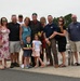 A group of approximately 12 people, including two young children, pose together on a military airfield tarmac. A man in a military flight suit stands at the center of the group, suggesting this may be a retirement ceremony, final flight, or homecoming event. A military aircraft is partially visible in the background.