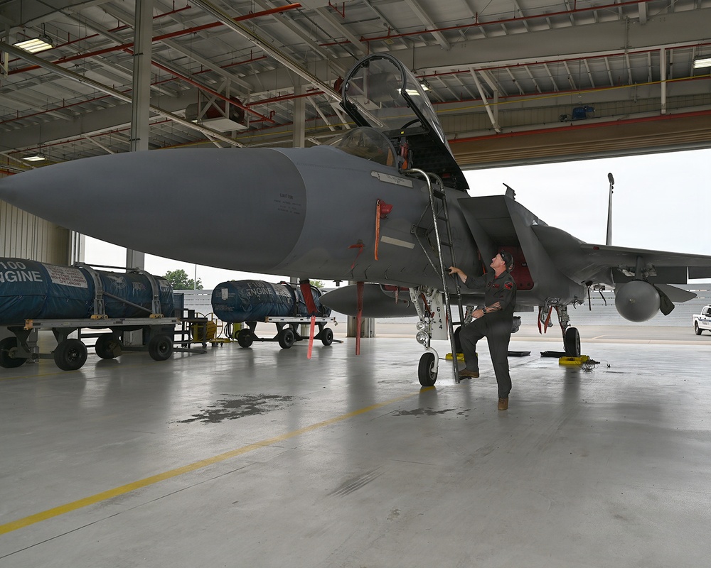 A military technician or pilot performs a pre-flight or maintenance inspection on an F-15 Eagle fighter jet inside a large aircraft hangar. The aircraft's canopy is open and the person is accessing the cockpit area while standing on the boarding ladder. Engine containers and ground support equipment are visible in the background.