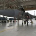 A military technician or pilot performs a pre-flight or maintenance inspection on an F-15 Eagle fighter jet inside a large aircraft hangar. The aircraft's canopy is open and the person is accessing the cockpit area while standing on the boarding ladder. Engine containers and ground support equipment are visible in the background.
