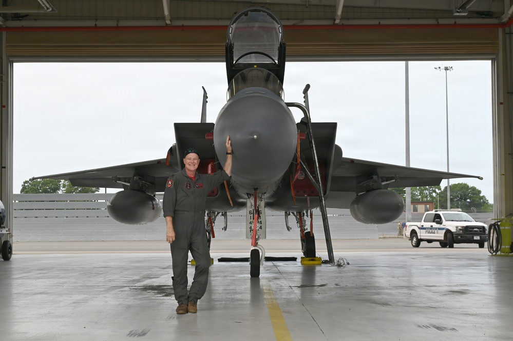 A military pilot in an olive green flight suit stands proudly in front of a large twin-engine fighter jet, likely an F-15 Eagle, inside a hangar bay. The aircraft's canopy is open and the jet is positioned facing forward, with external fuel tanks and weapons pylons visible. The setting appears to be a military air base with a police vehicle and personnel visible in the background.