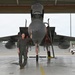 A military pilot in an olive green flight suit stands proudly in front of a large twin-engine fighter jet, likely an F-15 Eagle, inside a hangar bay. The aircraft's canopy is open and the jet is positioned facing forward, with external fuel tanks and weapons pylons visible. The setting appears to be a military air base with a police vehicle and personnel visible in the background.
