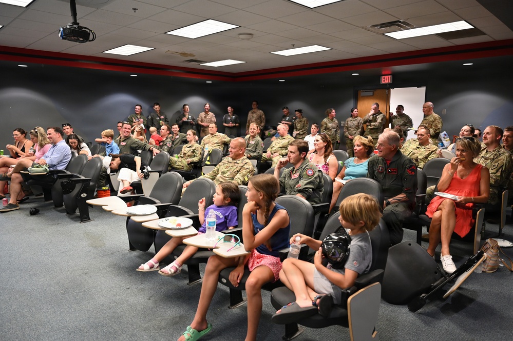 A large group of military personnel in camouflage uniforms and flight suits are gathered in what appears to be a briefing room or auditorium, accompanied by civilian family members including children. The event appears to be a military gathering or ceremony, possibly a homecoming, award ceremony, or unit function. The room features tiered seating with fold-out desk arms, fluorescent lighting, and dark walls with a red accent stripe.