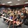 104th Fighter Wing Celebrates Flight Surgeon's Fifteen Years of Service with Fini Flight A large group of military personnel in camouflage uniforms and flight suits are gathered in what appears to be a briefing room or auditorium, accompanied by civilian family members including children. The event appears to be a military gathering or ceremony, possibly a homecoming, award ceremony, or unit function. The room features tiered seating with fold-out desk arms, fluorescent lighting, and dark walls with a red accent stripe.