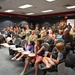 A large group of military personnel in camouflage uniforms and flight suits are gathered in what appears to be a briefing room or auditorium, accompanied by civilian family members including children. The event appears to be a military gathering or ceremony, possibly a homecoming, award ceremony, or unit function. The room features tiered seating with fold-out desk arms, fluorescent lighting, and dark walls with a red accent stripe.