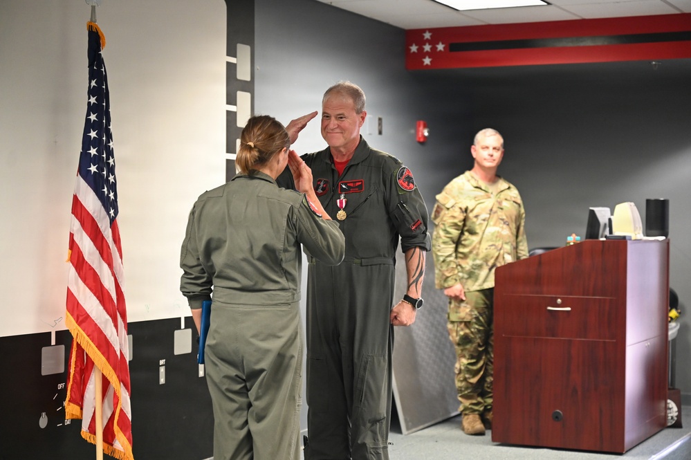 A military ceremony is taking place indoors where a female service member in olive green flight suit is saluting a male aviator who is wearing a flight suit with patches and a medal pinned to his chest. A third service member in camouflage uniform stands near a wooden podium in the background. An American flag is prominently displayed to the left.