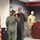 104th Fighter Wing Celebrates Flight Surgeon's Fifteen Years of Service with Fini Flight A military ceremony is taking place indoors where a female service member in olive green flight suit is saluting a male aviator who is wearing a flight suit with patches and a medal pinned to his chest. A third service member in camouflage uniform stands near a wooden podium in the background. An American flag is prominently displayed to the left.