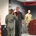 A military ceremony is taking place indoors where a female service member in olive green flight suit is saluting a male aviator who is wearing a flight suit with patches and a medal pinned to his chest. A third service member in camouflage uniform stands near a wooden podium in the background. An American flag is prominently displayed to the left.