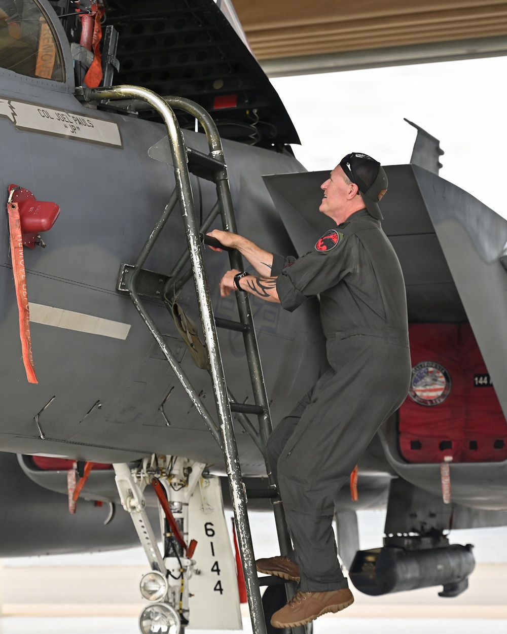 A person in an olive/sage green flight suit is climbing a boarding ladder to access the cockpit of a military jet aircraft, likely an F-15. The aircraft is gray and bears a name placard reading 'COL JOEL HAULS' with a callsign 'JP', along with a squadron patch visible on the fuselage. The scene appears to be inside or just outside a hangar.