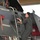 104th Fighter Wing Celebrates Flight Surgeon's Fifteen Years of Service with Fini Flight A person in an olive/sage green flight suit is climbing a boarding ladder to access the cockpit of a military jet aircraft, likely an F-15. The aircraft is gray and bears a name placard reading 'COL JOEL HAULS' with a callsign 'JP', along with a squadron patch visible on the fuselage. The scene appears to be inside or just outside a hangar.