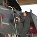 A person in an olive/sage green flight suit is climbing a boarding ladder to access the cockpit of a military jet aircraft, likely an F-15. The aircraft is gray and bears a name placard reading 'COL JOEL HAULS' with a callsign 'JP', along with a squadron patch visible on the fuselage. The scene appears to be inside or just outside a hangar.