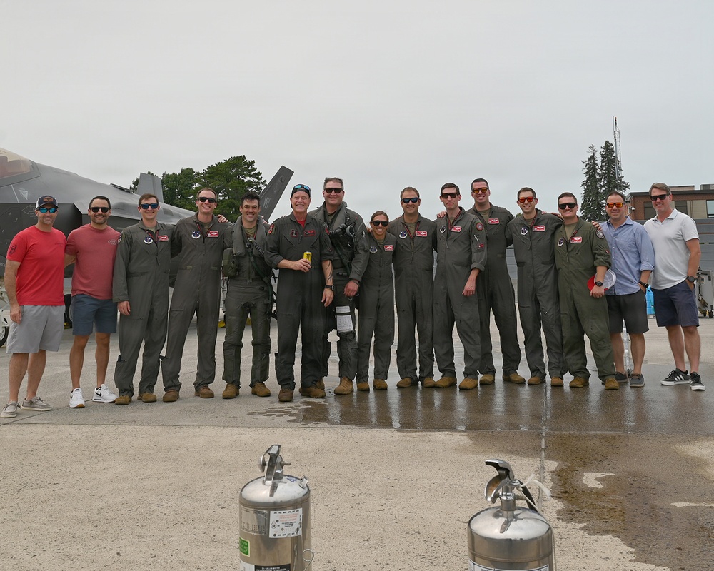 A group of approximately 14 individuals pose for a group photo on a wet aircraft ramp or tarmac, with at least one military jet aircraft (appearing to be an F-35) visible in the background. The majority of the group are wearing olive/sage green flight suits consistent with U.S. Air Force attire, while a few individuals on the flanks are in civilian clothing. The setting appears to be a military air base or airshow flight line.