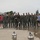 104th Fighter Wing Celebrates Flight Surgeon's Fifteen Years of Service with Fini Flight A group of approximately 14 individuals pose for a group photo on a wet aircraft ramp or tarmac, with at least one military jet aircraft (appearing to be an F-35) visible in the background. The majority of the group are wearing olive/sage green flight suits consistent with U.S. Air Force attire, while a few individuals on the flanks are in civilian clothing. The setting appears to be a military air base or airshow flight line.