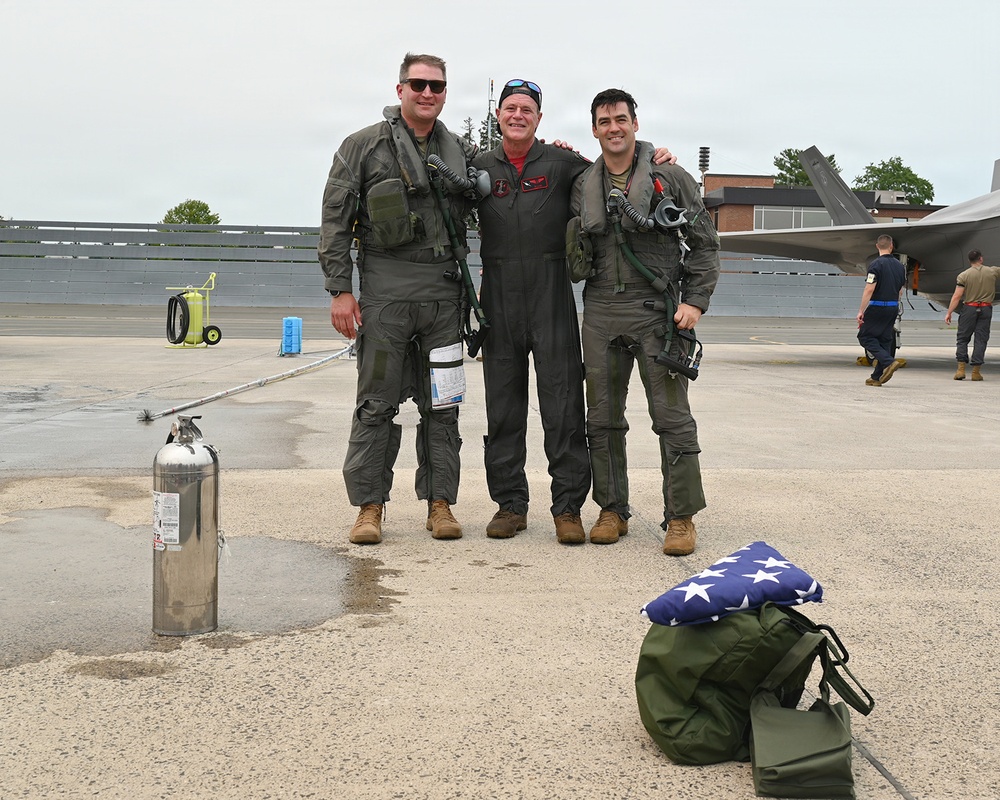 Three individuals in military flight suits pose together on an aircraft flight line or tarmac. Two of the three are wearing full flight gear including G-suits, survival vests, and oxygen masks, while the center individual wears a standard flight suit. A folded American flag and military gear bag are visible in the foreground, suggesting a ceremonial or milestone occasion.