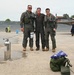 Three individuals in military flight suits pose together on an aircraft flight line or tarmac. Two of the three are wearing full flight gear including G-suits, survival vests, and oxygen masks, while the center individual wears a standard flight suit. A folded American flag and military gear bag are visible in the foreground, suggesting a ceremonial or milestone occasion.