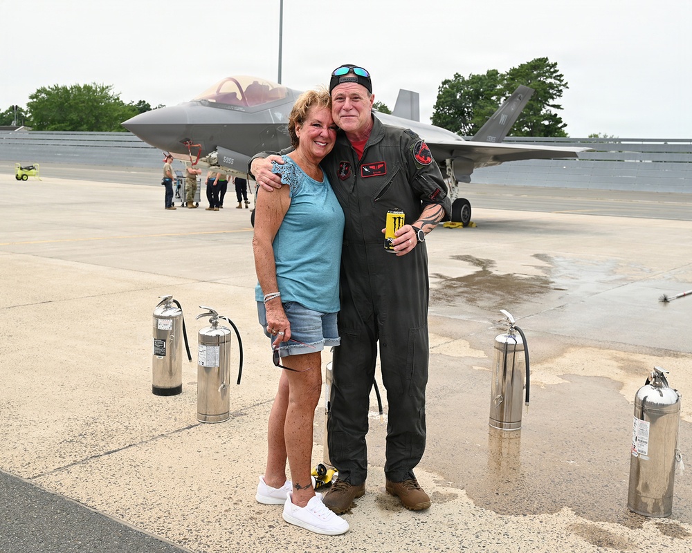 A military pilot in a flight suit poses with a woman on an aircraft tarmac in front of an F-35 fighter jet. The pilot has his arm around the woman and is holding a Monster Energy drink. The setting appears to be a military air base, likely during a post-flight celebration or reunion.