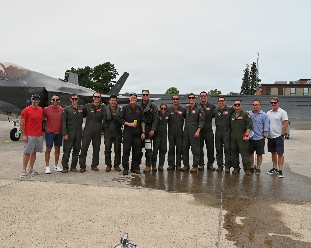 A group of approximately 14 individuals pose for a group photo on a wet aircraft ramp in front of what appears to be an F-35 fighter jet. The majority of the group is wearing olive/sage green military flight suits, while two individuals on the far left and two on the far right are dressed in civilian clothing. The setting appears to be a military air base or airshow flight line.