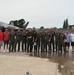 A group of approximately 14 individuals pose for a group photo on a wet aircraft ramp in front of what appears to be an F-35 fighter jet. The majority of the group is wearing olive/sage green military flight suits, while two individuals on the far left and two on the far right are dressed in civilian clothing. The setting appears to be a military air base or airshow flight line.